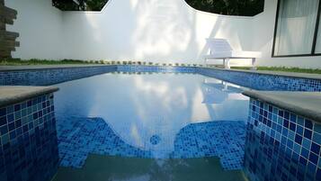 Piscina com azulejos azuis, rodeada por um muro branco com uma janela circular e uma pequena área de estar ao ar livre.