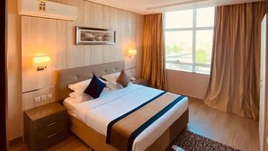 A hotel room with a bed, bedside tables, a nightstand, a window with curtains, and a wall-mounted air conditioning unit. - Banan Hotel Suite (Tabuk)