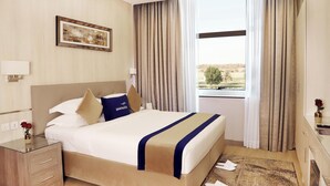 A hotel room with a bed, a nightstand, a desk, a window with curtains, and a painting on the wall. - Banan Hotel Suite (Tabuk)