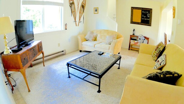 Una sala de estar con un sofá, una mesa de centro, un televisor sobre un mueble de madera y una ventana con cortinas.