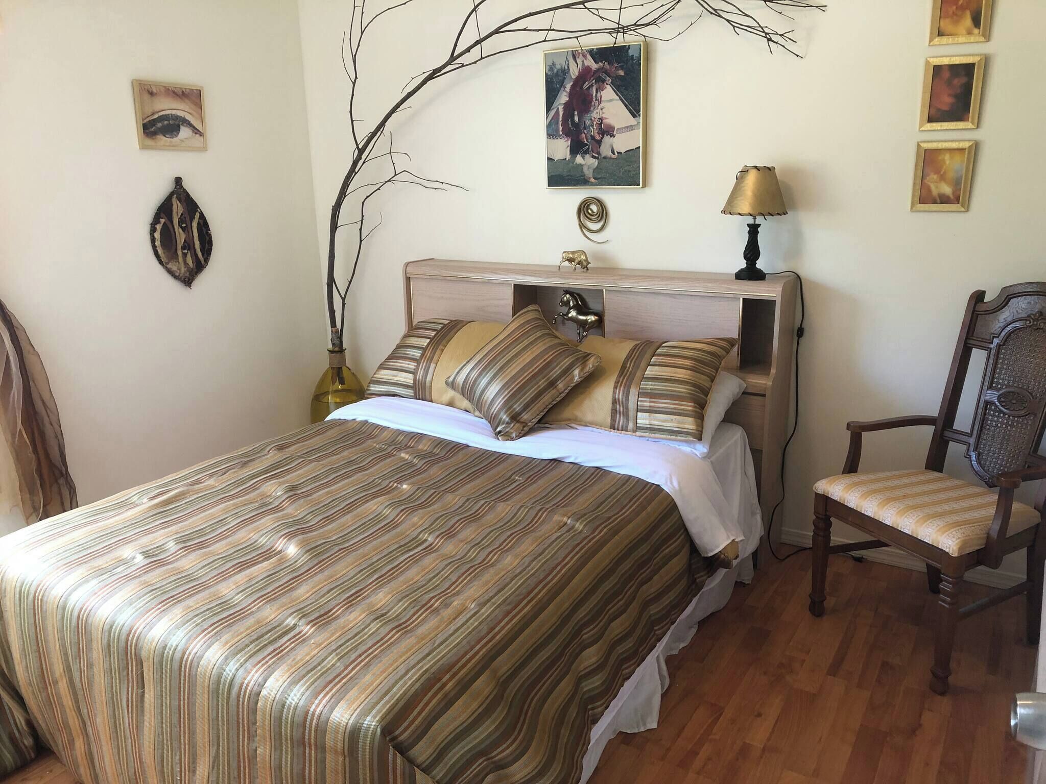 A bedroom with a bed, a chair, a lamp, and wall decorations including a picture of a person and a tree branch.