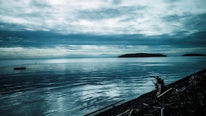 Una masa de agua tranquila con una isla distante, un muelle y una costa rocosa.