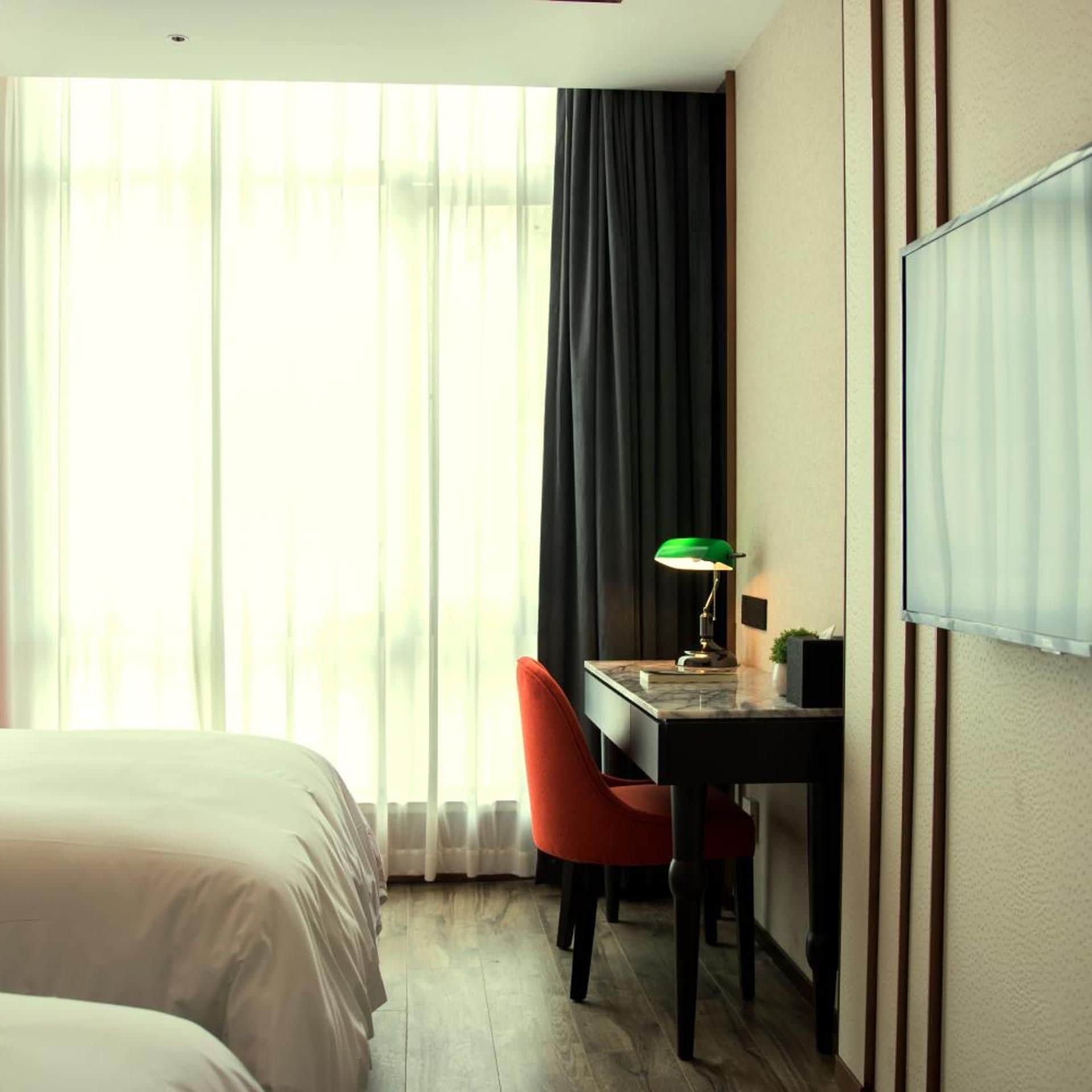 Une chambre d’hôtel avec un lit, un bureau doté d’une lampe, une chaise et une grande fenêtre avec des rideaux.