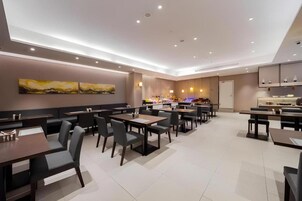 A modern restaurant with multiple dining tables and chairs, a kitchen area in the background, and artwork on the walls.