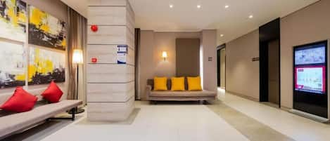 A hotel lobby with a reception desk, a sofa with yellow cushions, a wall-mounted TV, and a ceiling with recessed lighting.