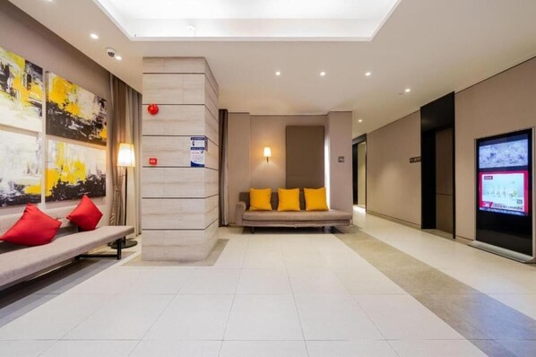 Un vestíbulo de hotel con un mostrador de recepción, un sofá con cojines amarillos, un televisor montado en la pared e iluminación empotrada en el techo.