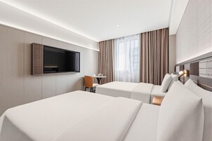 Habitación de hotel con dos camas, un televisor montado en la pared, una mesita con una silla y una ventana con cortinas.