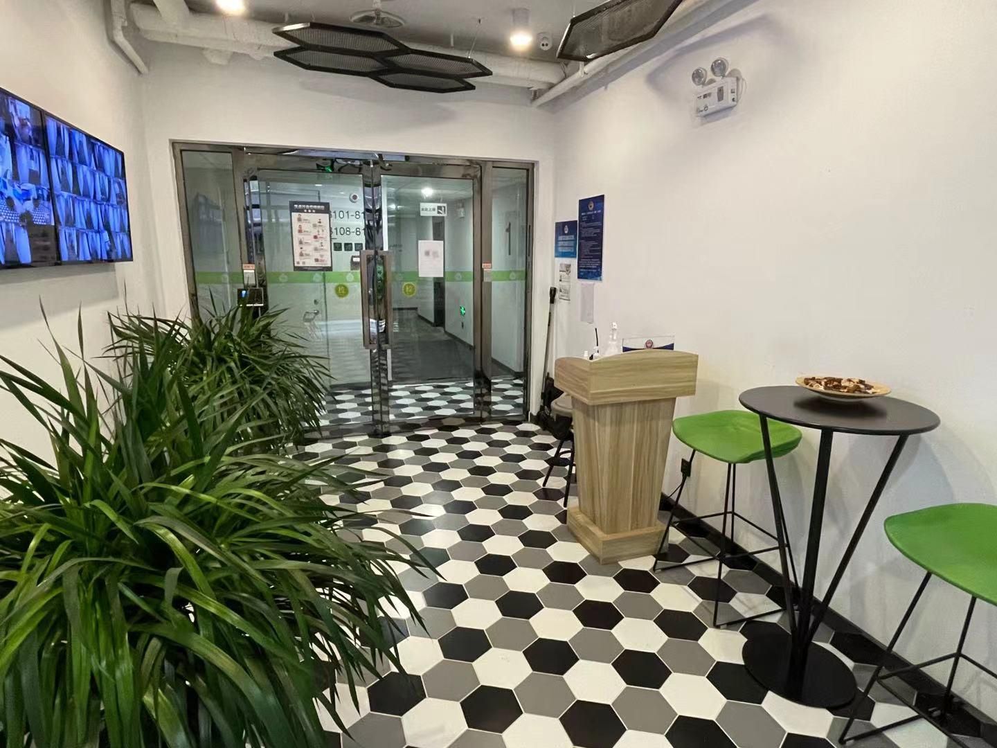 Zona de recepción con piso a cuadros, un atril de madera, sillas verdes y una planta en maceta.