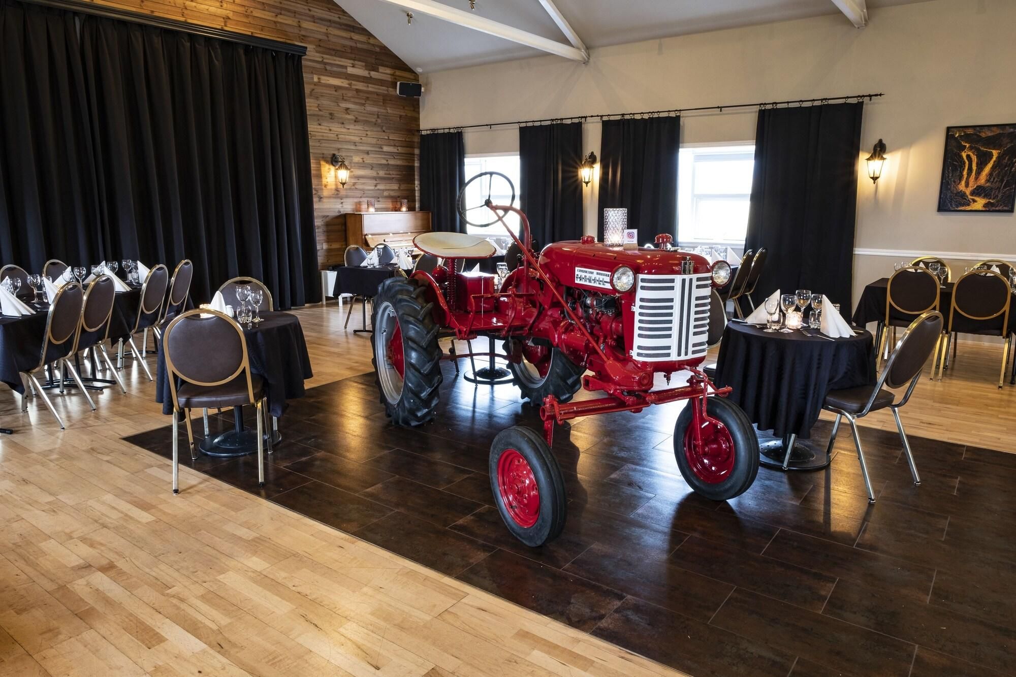 Un espace repas avec des tables dressées pour le repas, un tracteur rouge au centre et un mur en bois avec une fenêtre.