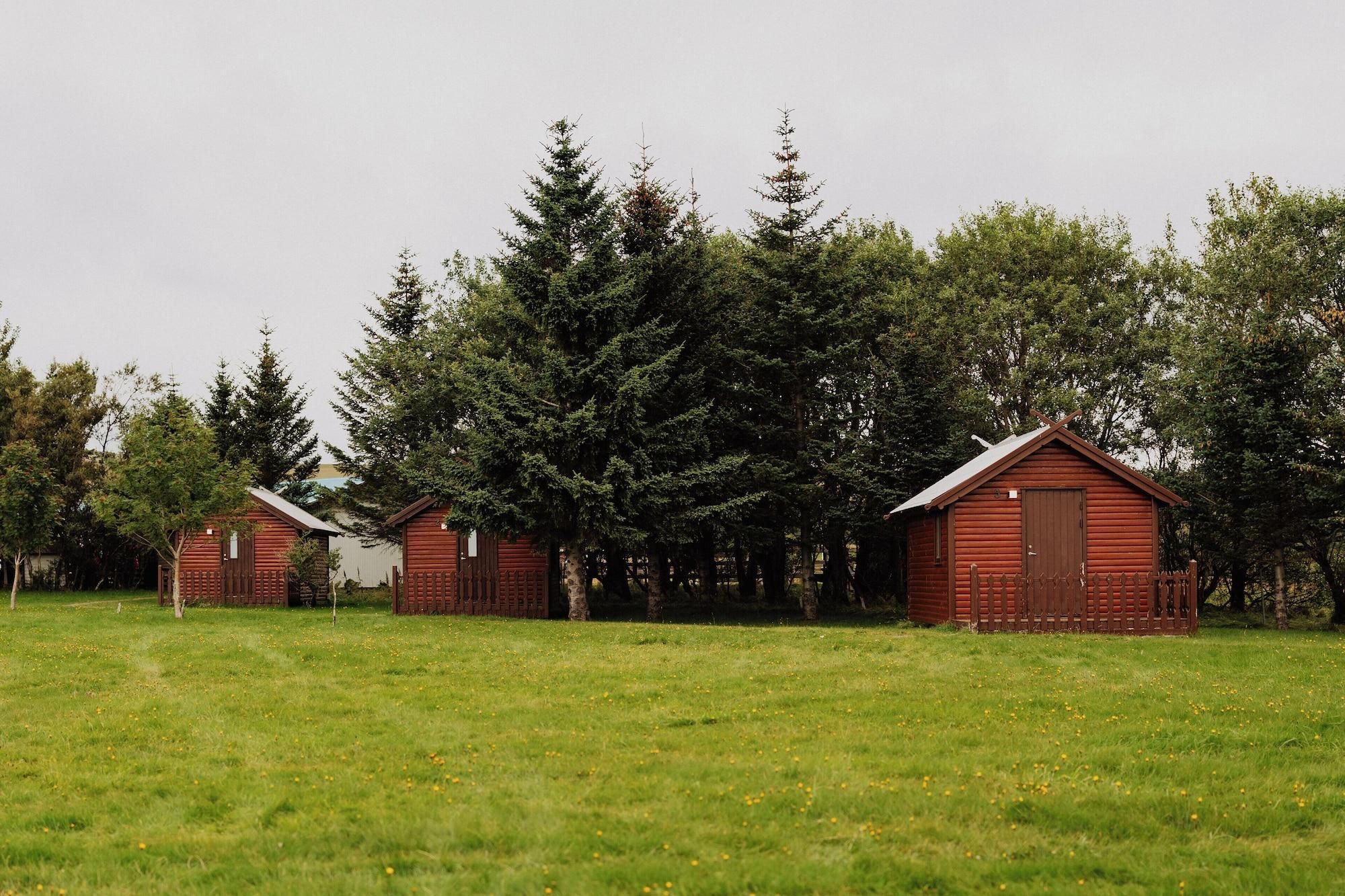 Une rangée de cabanes en bois rouge se dresse dans un champ verdoyant, avec une forêt dense en arrière-plan.