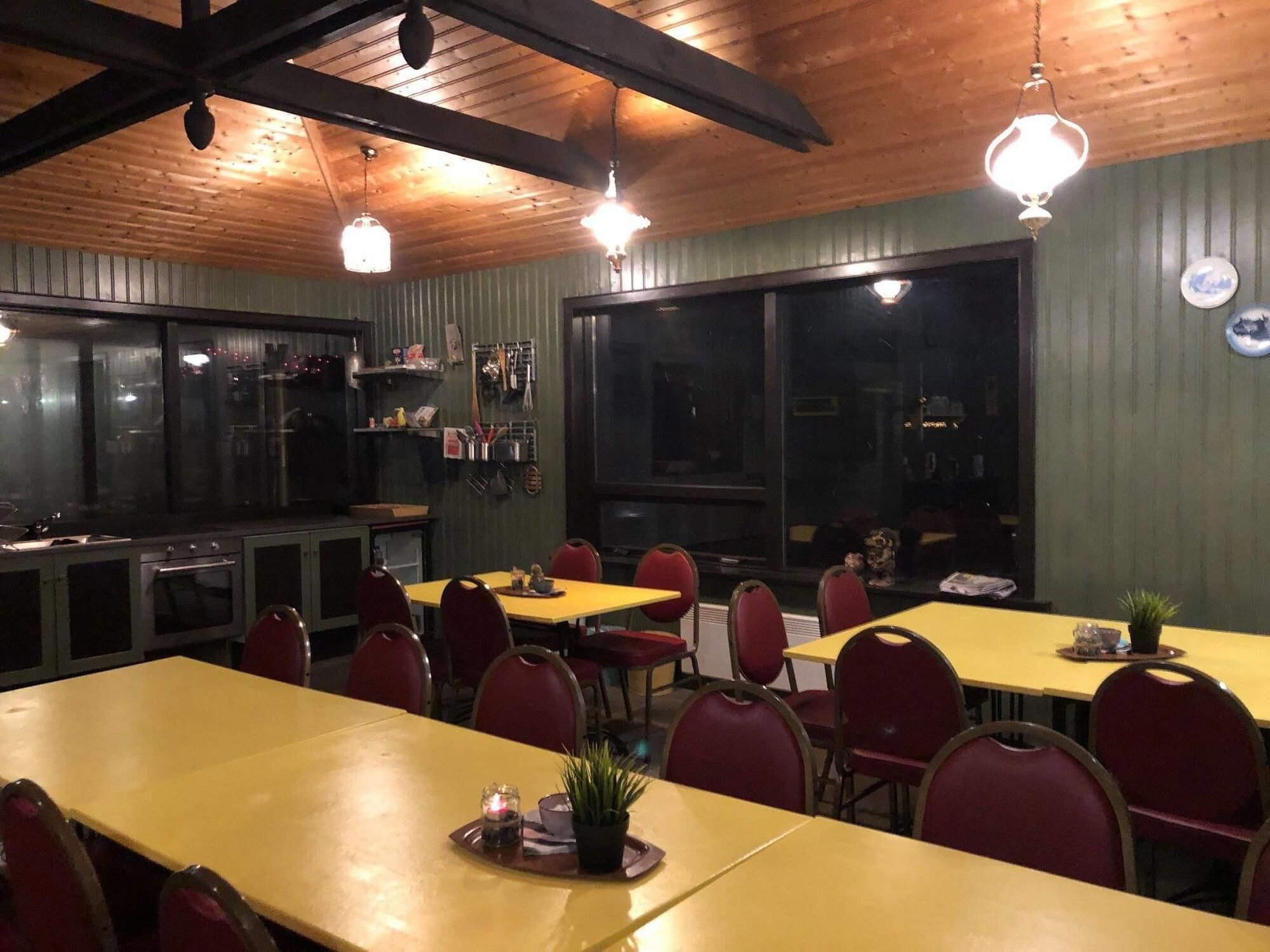 A restaurant with wooden ceiling beams, pendant lighting, and a large window. Tables are set with red chairs and centerpieces.
