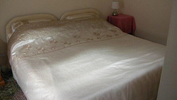 A bed with a floral-patterned bedspread, a bedside table with a lamp, and a small red tablecloth. - Watermill Farm (Lincoln)