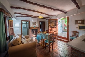 Un salon chaleureux avec une cheminée, un coin repas avec une table dressée pour deux personnes, et une vue sur le jardin par une porte ouverte.