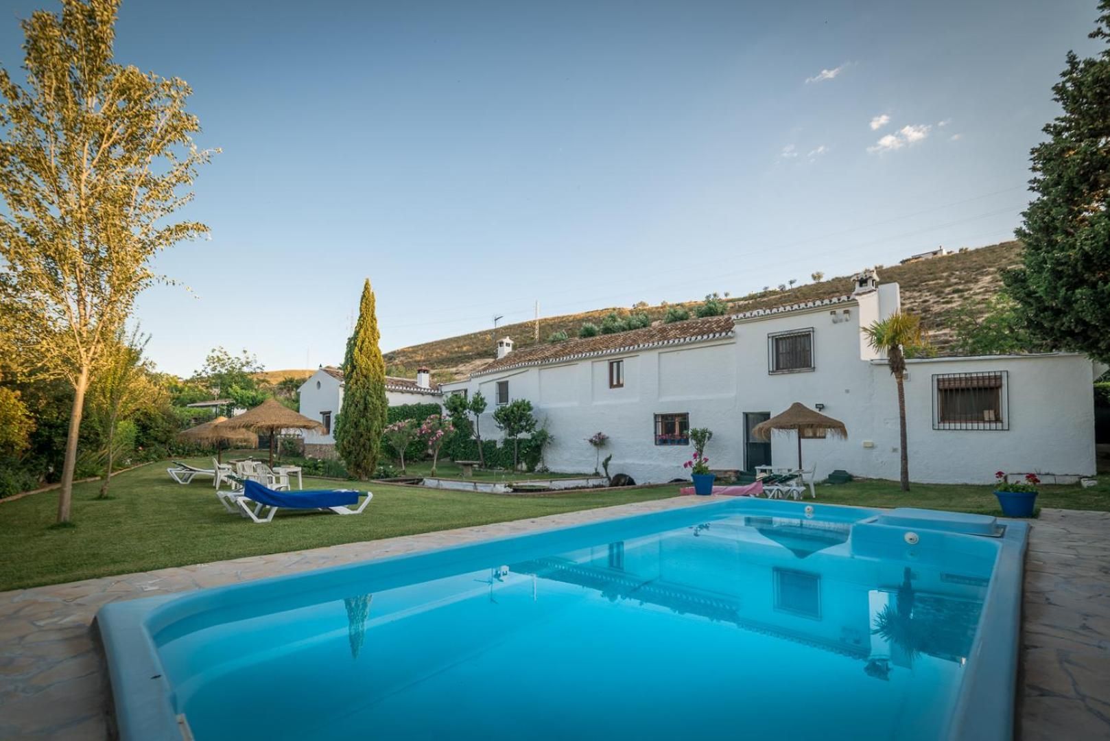 A swimming pool with lounge chairs, a white building with a tiled roof, and a grassy area with trees.
