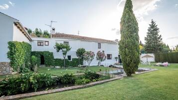 A white building with a tiled roof, surrounded by a well-maintained garden with various plants and a small pond.