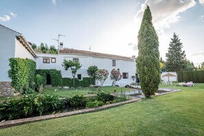 Un bâtiment blanc, doté d’un toit recouvert de tuiles, entouré d’un jardin bien entretenu où poussent différentes espèces de plantes et où se trouve un petit étang.
