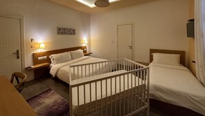 A hotel room with two beds, a crib, a wooden headboard, a nightstand, a wall-mounted lamp, and a door. - Hotel Green House (Berat)