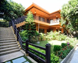 Un edificio de dos pisos con fachada de madera, una escalera con barandilla negra y un jardín con diversas plantas y flores.