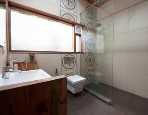 Un baño con tocador de madera, lavamanos blanco, cabina de ducha de vidrio y baldosas estampadas.
