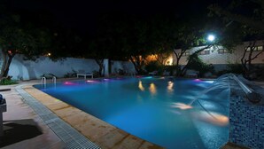 A swimming pool at night with a water feature, surrounded by trees and a building with illuminated windows.