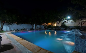 A swimming pool at night with a water feature, surrounded by trees and a building with illuminated windows.