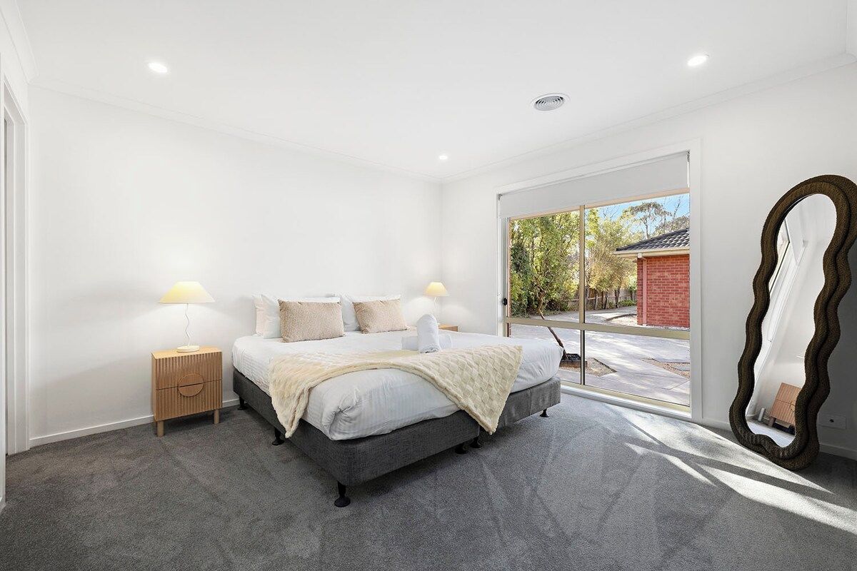 A bedroom with a bed, bedside lamps, a large mirror, and a view through a window.