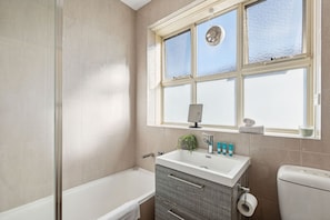A bathroom with a bathtub, a sink with toiletries, a window with a view, and a toilet. - Domi Rentals - The Carlisle Street Apartments (Balaclava)