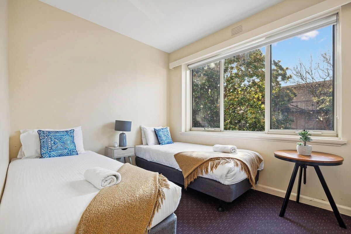 A hotel room with two beds, a window with a view of trees, a small table with a plant, and a nightstand with a lamp.