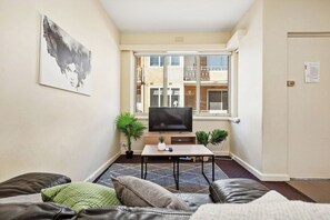 A compact living space with a sofa, a television, and a window with a view of a building. - Domi Rentals - The Carlisle Street Apartments (Balaclava)
