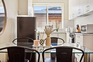 A modern kitchen with white cabinets, a glass dining table, and a view of a brick building outside. - Domi Rentals - The Carlisle Street Apartments (Balaclava)