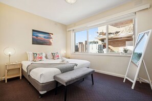 A bedroom with a large bed, a bench, a bedside table, a floor lamp, a painting, and a window with a view of buildings. - Domi Rentals - The Carlisle Street Apartments (Balaclava)