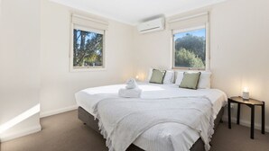 A bedroom with a bed, two windows, an air conditioning unit, and a bedside table with a lamp. - Domi Rentals The Blackburn Residence (Notting Hill)