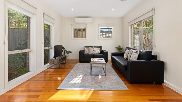A spacious living room with hardwood flooring, two black sofas, a coffee table, and large windows with views of greenery. - Domi Rentals The Blackburn Residence (Notting Hill)