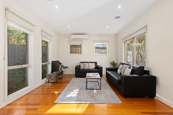 A spacious living room with hardwood flooring, two black sofas, a coffee table, and large windows with views of greenery.