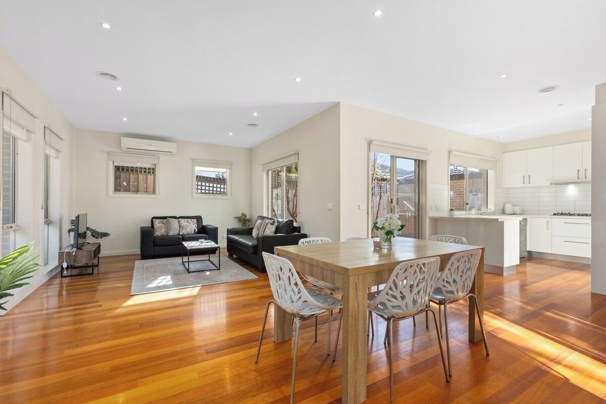 A spacious living area with a wooden dining table, white chairs, a black sofa, and a kitchen area with white cabinets.