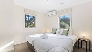 A bedroom with a bed, two windows, an air conditioning unit, and a bedside table with a lamp.