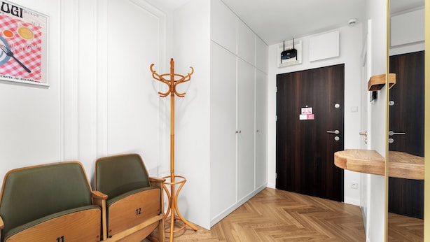 Una habitación con dos sillas verdes, un perchero, un banco de madera, una puerta oscura y una pared blanca con un cartel.