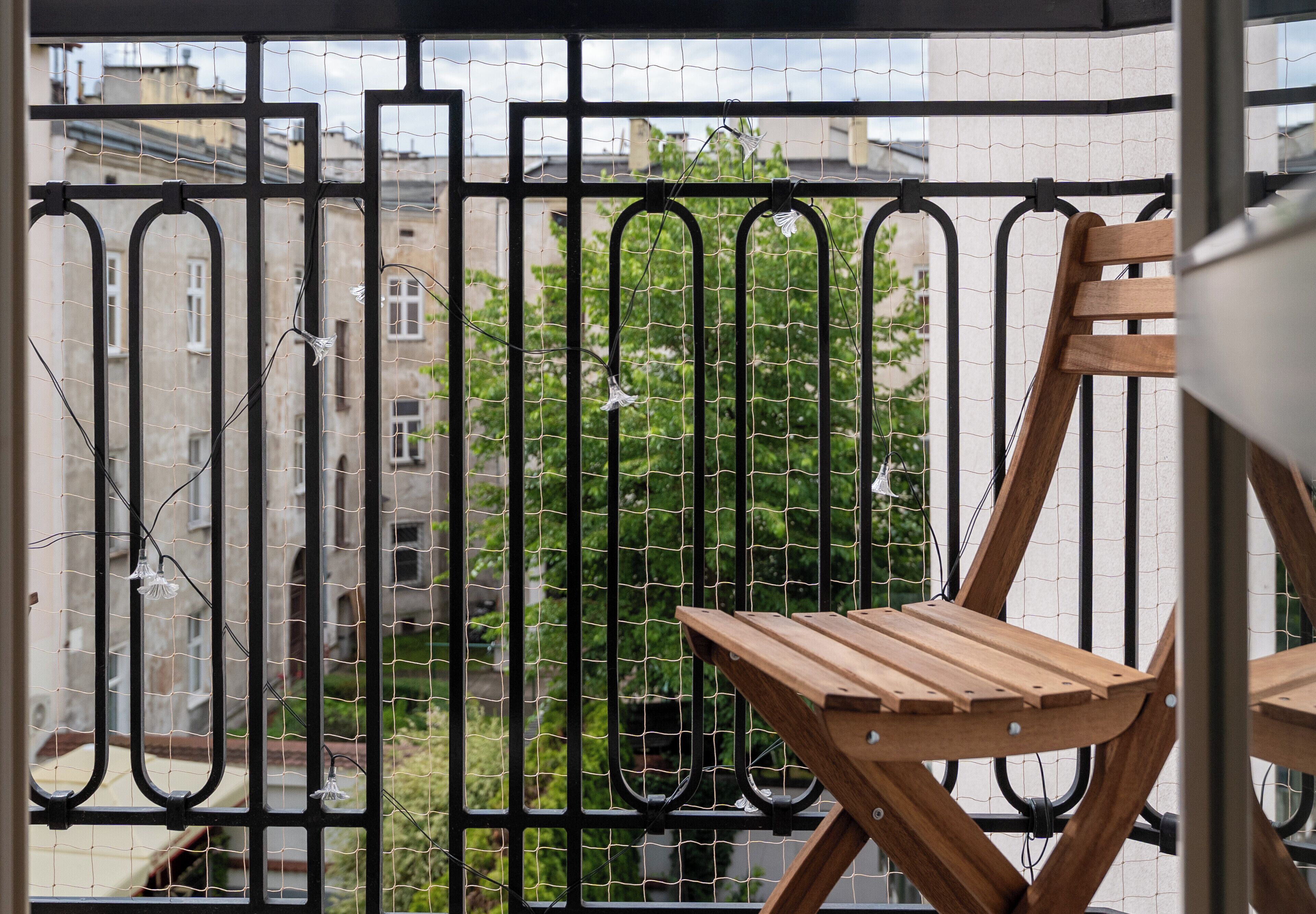 Un balcon avec une chaise en bois, une rambarde en métal et une vue sur des bâtiments et de la verdure.