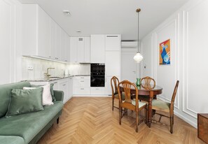 A modern kitchen with white cabinetry, a dining table with chairs, and a green sofa. - Kamienica Pod Czaplą Heritage Apartment (Kraków)