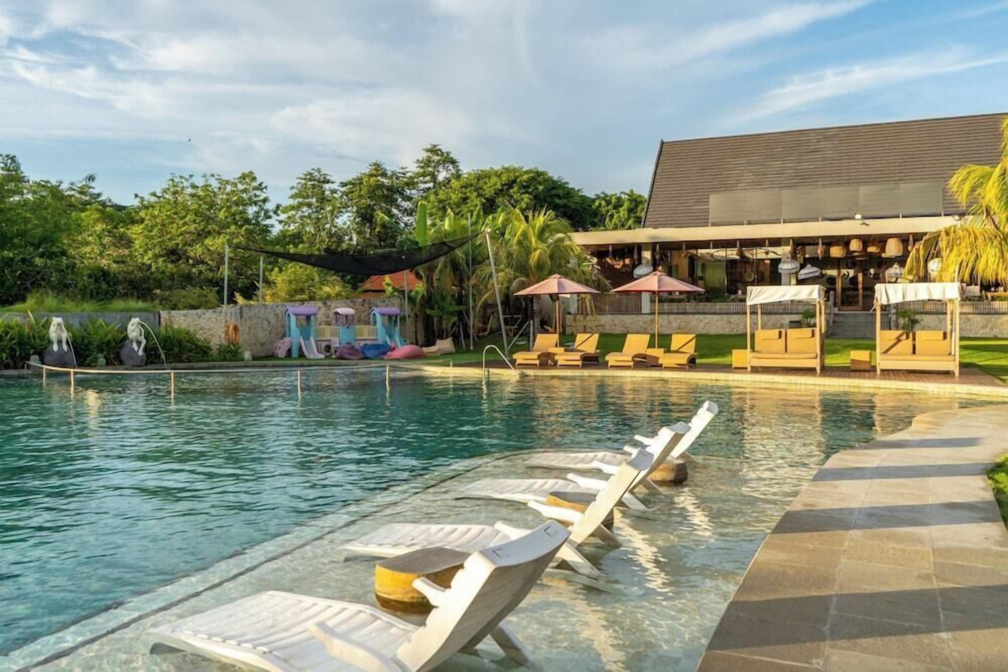 A swimming pool with lounge chairs and umbrellas, surrounded by greenery and a building with a sloped roof.