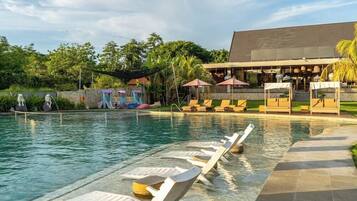A swimming pool with lounge chairs and umbrellas, surrounded by greenery and a building with a sloped roof.
