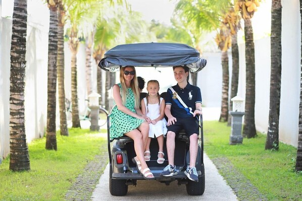 Une famille de trois personnes se promène en voiturette de golf sur un chemin bordé de palmiers.