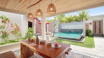 A wooden dining table with hanging lights, a pool, and a garden with pink flowers.
