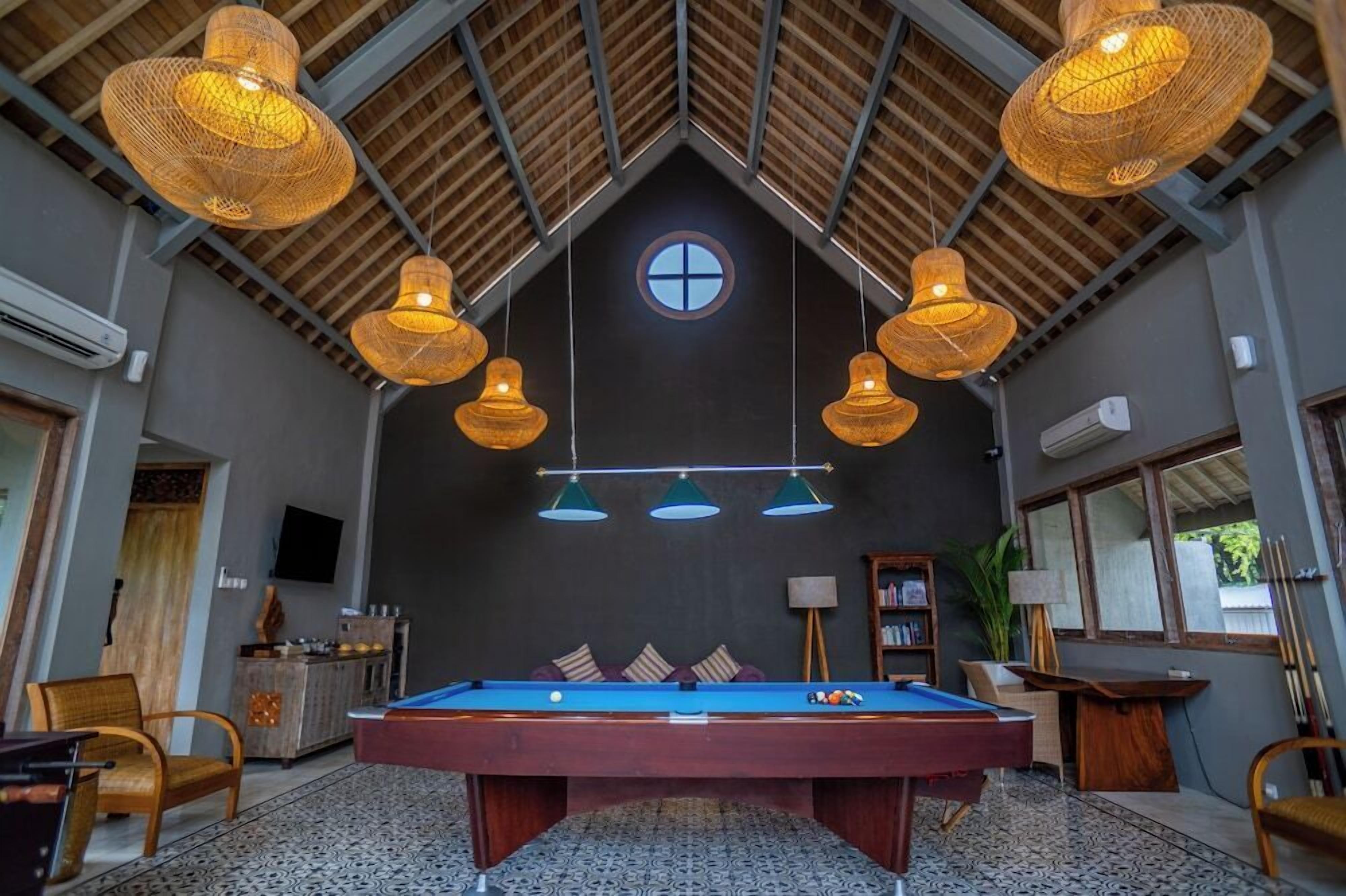 A pool table in a room with a high ceiling, hanging wicker lamps, and a television on the wall.