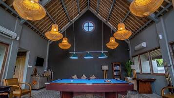 A pool table in a room with a high ceiling, hanging wicker lamps, and a television on the wall.