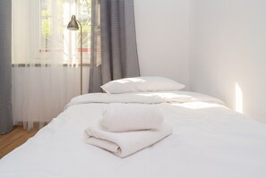 Cama arrumada com almofadas e toalha dobrada, abajur e cortinas leves com vista para o verde lá fora.