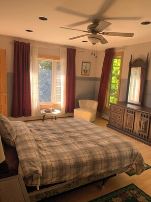 Ein Schlafzimmer mit Bett, Sessel, Kommode, Deckenventilator und einem Fenster mit Vorhängen.
