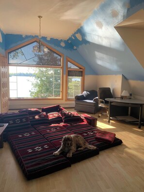 Una habitación con un ventanal grande, techo con vigas a la vista, sofá con cojines estampados, un perro pequeño y un escritorio con lámpara.