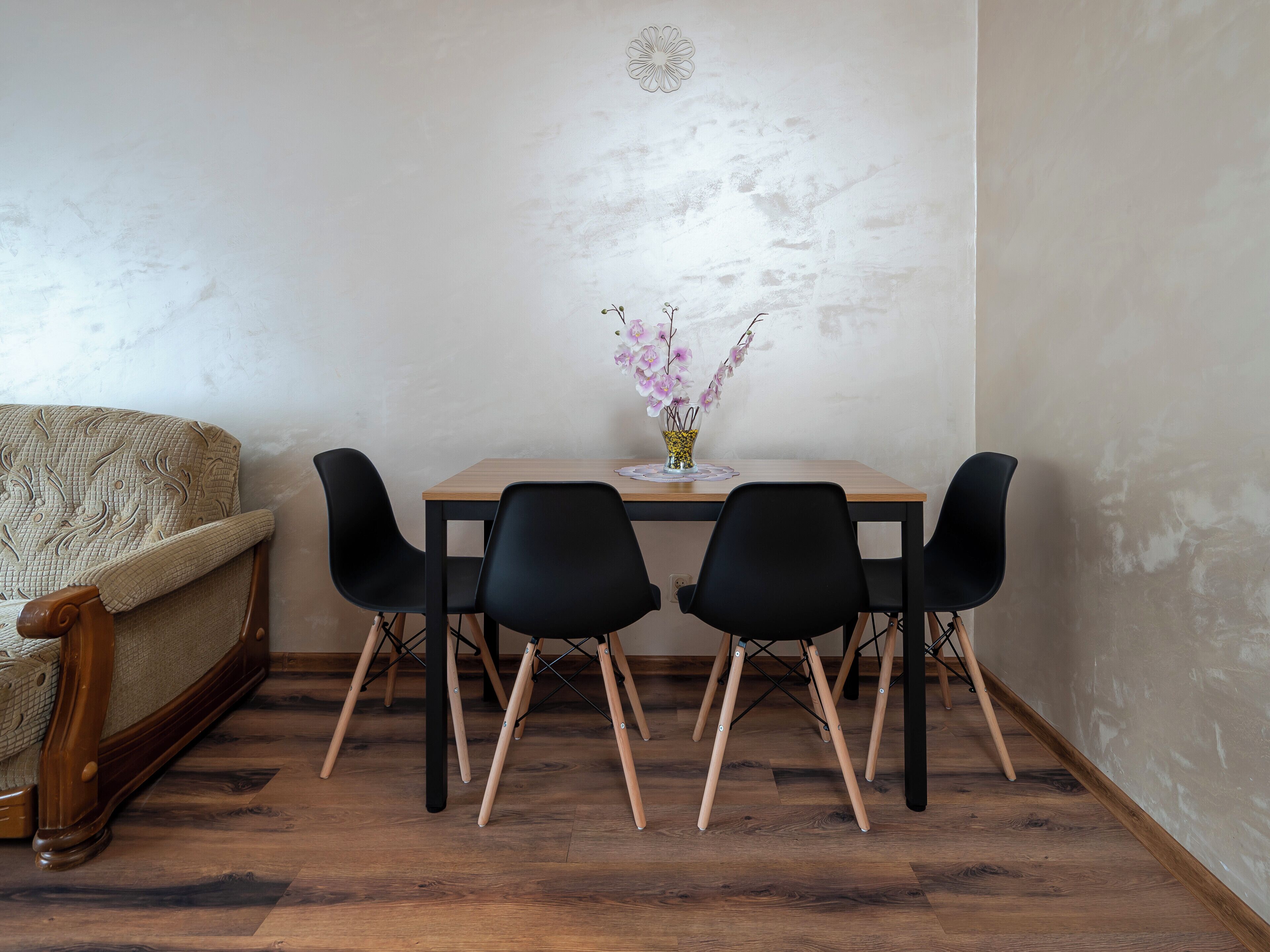 A dining area with a wooden table, four black chairs, a vase with flowers, and a sofa.