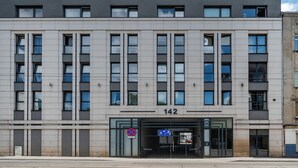 A modern building with a flat facade, numbered 142, featuring a ground-level entrance with a glass door and a pedestrian crossing sign. - Park Źródliska Harmony Studio (Łódź)